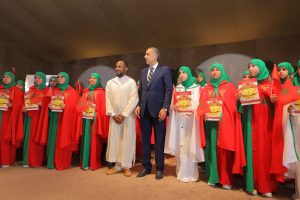 Group of women in green hijabs and red coats holding posters and Moroccan flags at a ceremony, flanked by two men in the center.