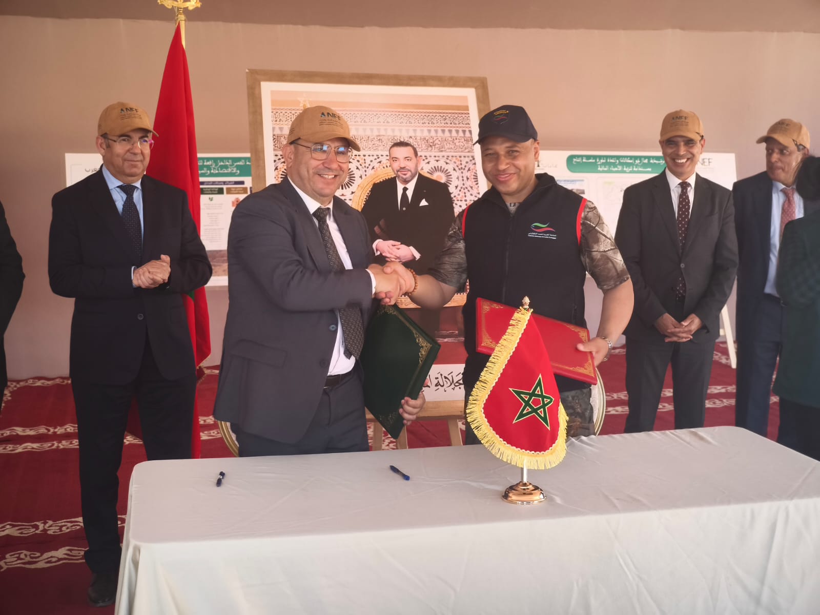 Two men in suits shake hands over a table during a formal ceremony, with a Moroccan flag on the table and several officials watching in the background.