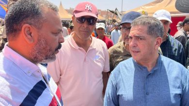 Three men converse at an outdoor event with tents and a crowd in the background; the man on the left wears a striped shirt, the middle man in a pink polo and NY cap, and the man on the right in a blue traditional shirt.