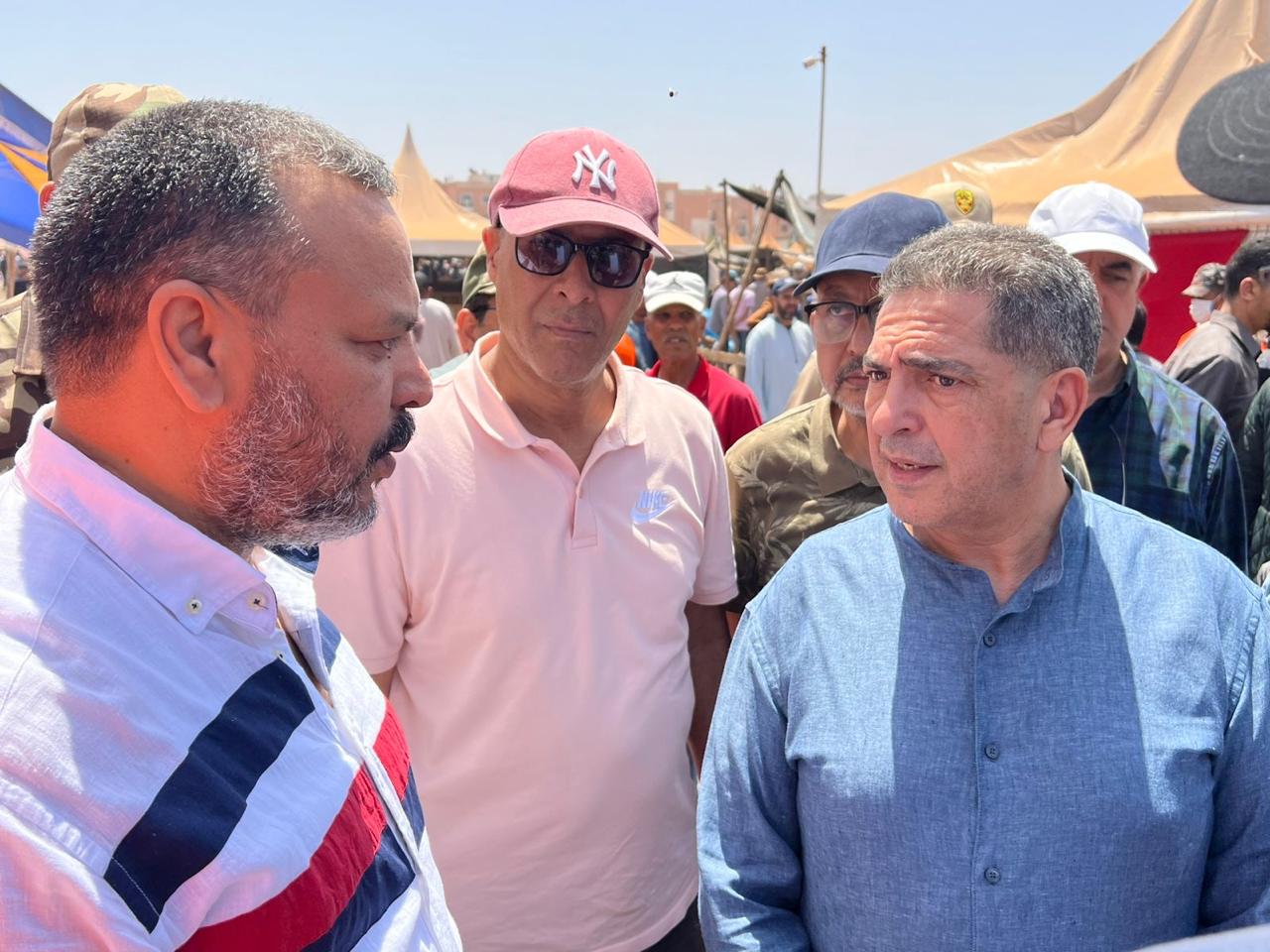 IMG-20260523-WA01144 | MCG24 | موقع إلكتروني مغربي شامل متجدد على مدار الساعة Three men converse at an outdoor event with tents and a crowd in the background; the man on the left wears a striped shirt, the middle man in a pink polo and NY cap, and the man on the right in a blue traditional shirt.