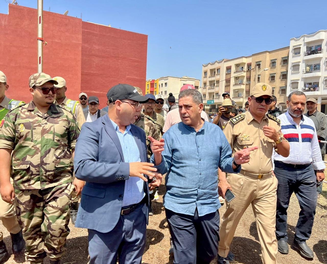 Group of officials and soldiers walking and talking in a sunny urban street, with colorful buildings in the background.