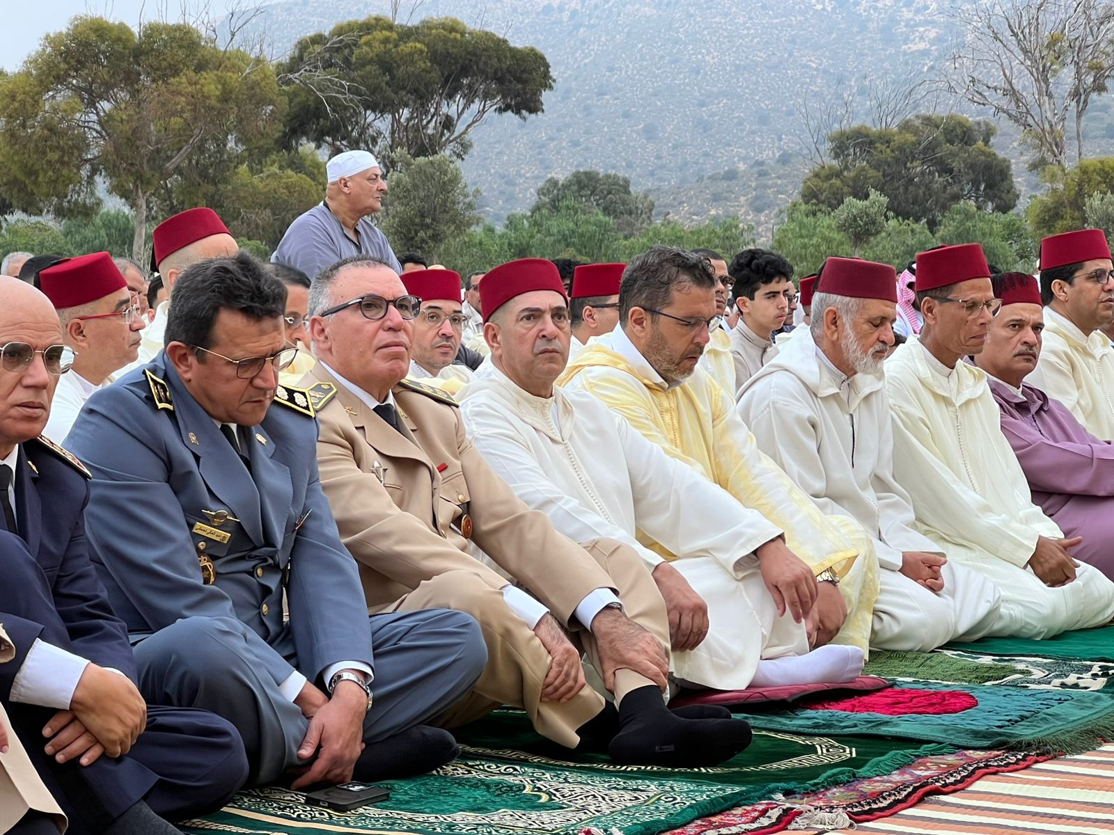 Group of men in traditional Moroccan clothing and uniforms seated on prayer rugs at an outdoor ceremony with mountains in the background.