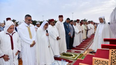 Group of men in white traditional robes standing in prayer on red carpets during an outdoor religious ceremony; a man holds a microphone.