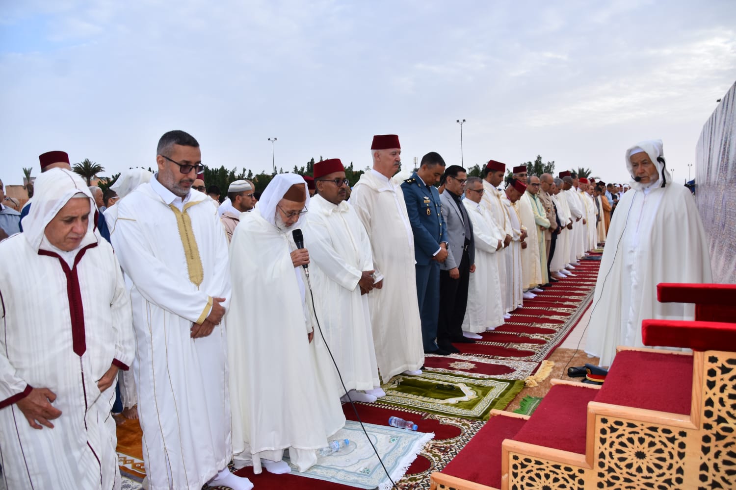 Group of men in white traditional robes standing in prayer on red carpets during an outdoor religious ceremony; a man holds a microphone.