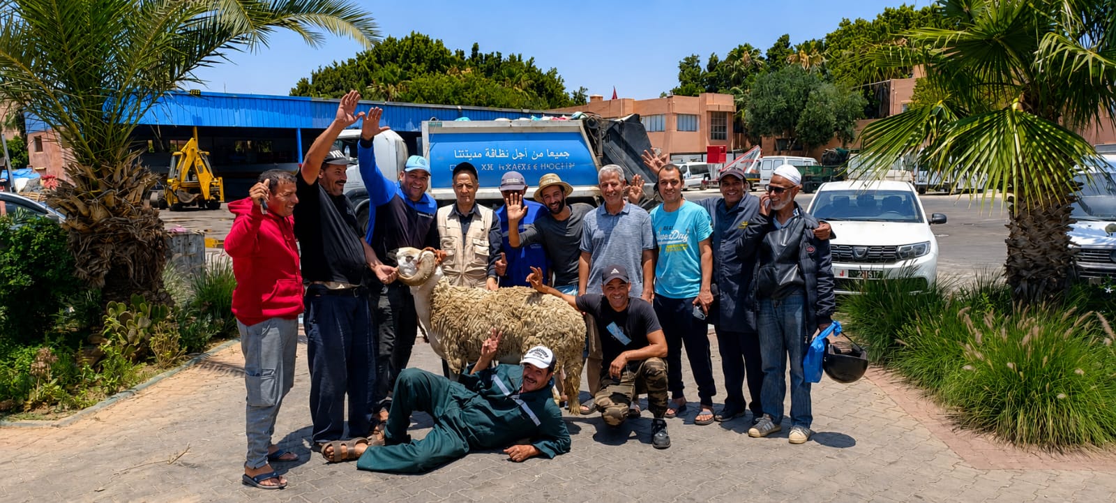 IMG-20260531-WA0193 | MCG24 | موقع إلكتروني مغربي شامل متجدد على مدار الساعة Group of men posing with a sheep in a sunny outdoor lot, with trucks and palm trees in the background.