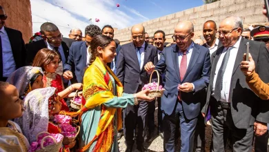 In a sunny outdoor scene, formally dressed officials in suits stand with smiling children who present flower baskets and wear bright traditional clothing.