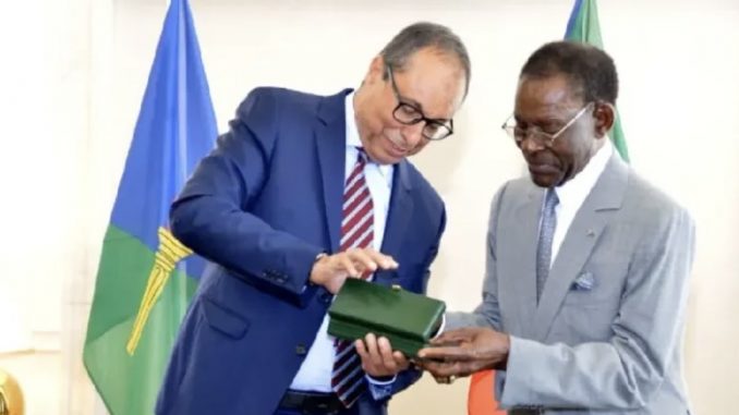 Two men in suits exchange a green box during a formal ceremony, with flags in the background.
