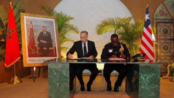 IMG_1901 | MCG24 | موقع إلكتروني مغربي شامل متجدد على مدار الساعة Two men in suits sign documents at a marble desk during a formal signing ceremony, flanked by Moroccan and U.S. flags and a framed portrait nearby.