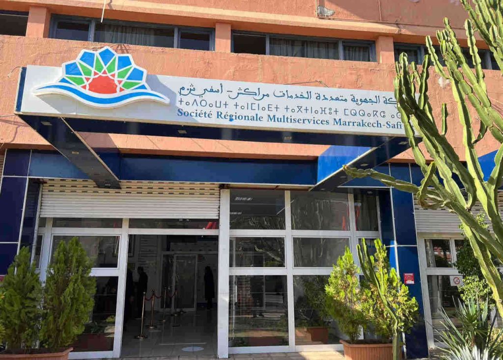 IMG_2794 | MCG24 | موقع إلكتروني مغربي شامل متجدد على مدار الساعة Front entrance of the Société Régionale Multiservices Marrakech-Safi office with a colorful logo sign and glass doors, potted cacti outside.