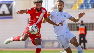 Two soccer players compete for the ball during a match; one in red kit, the other in light blue.