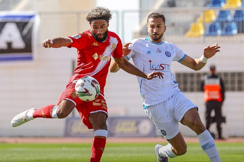 Two soccer players compete for the ball during a match; one in red kit, the other in light blue.