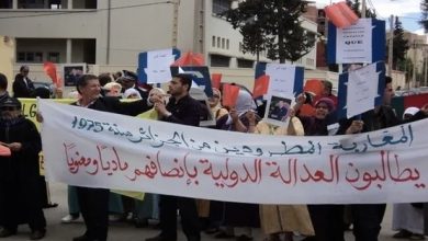 Protesters march on a street, holding a long Arabic banner and various placards and red/blue signs behind them.