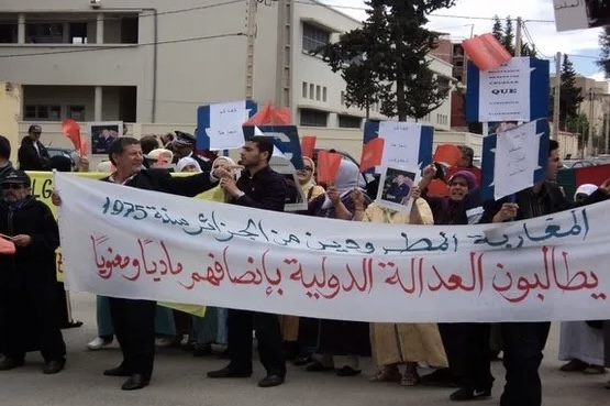Protesters march on a street, holding a long Arabic banner and various placards and red/blue signs behind them.
