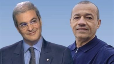 Two men pose together against a blue gradient background; one in a suit and tie, the other in a dark polo.