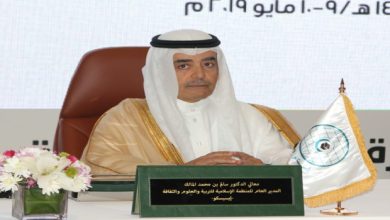 Arab man in a white thobe and ghutra sits at a conference table with a green nameplate, flower arrangement, and a flag with a blue emblem.
