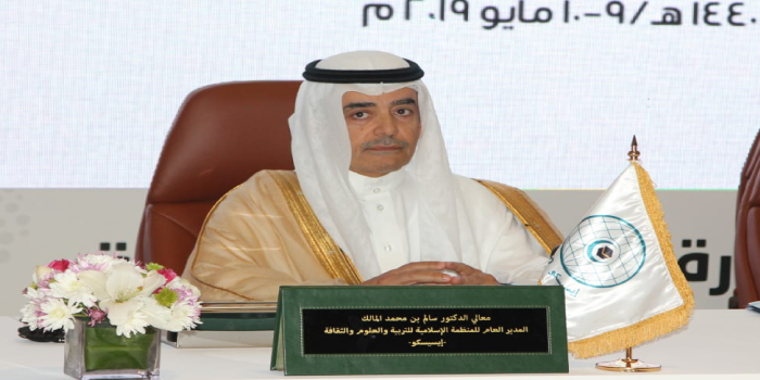 Arab man in a white thobe and ghutra sits at a conference table with a green nameplate, flower arrangement, and a flag with a blue emblem.