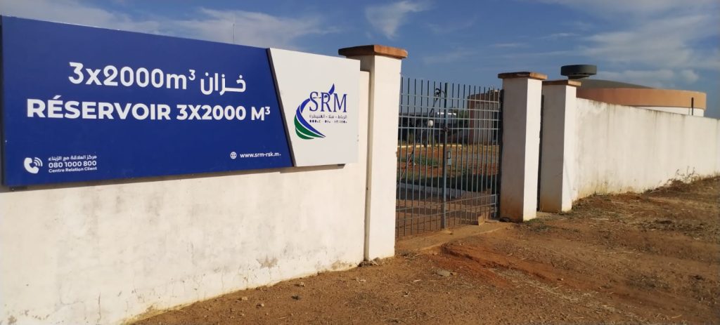 Blue sign on a white wall advertising a 3x2000 m³ reservoir, with SRM logo; gate and fencing visible behind.
