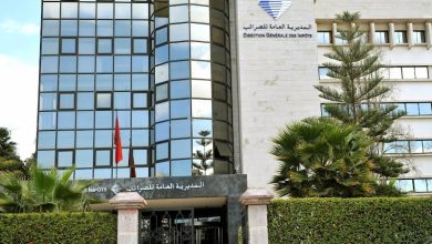 Front of a government tax office building with a glass facade and bilingual Arabic/French signage, framed by palm trees.