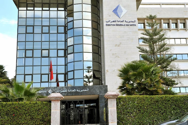 Front of a government tax office building with a glass facade and bilingual Arabic/French signage, framed by palm trees.