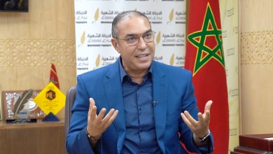 Man in a blue suit speaking and gesturing with his hands during an interview or talk, seated in a studio with a Moroccan flag in the background and a patterned wall behind him.