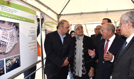 Group of officials in suits examine an architectural display board with Arabic text and photos inside a tent.