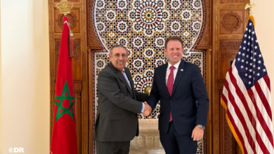 Two men in business suits shake hands in front of a decorative mosaic wall, flanked by the Moroccan flag on the left and the U.S. flag on the right.