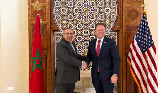 Two men in business suits shake hands in front of a decorative mosaic wall, flanked by the Moroccan flag on the left and the U.S. flag on the right.