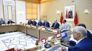 MAP021798798 | MCG24 | موقع إلكتروني مغربي شامل متجدد على مدار الساعة Formal government meeting with officials seated along a long ornate table in a wood-paneled room, Moroccan flags and a portrait on the wall.