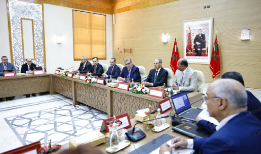 MAP021798798 | MCG24 | موقع إلكتروني مغربي شامل متجدد على مدار الساعة Formal government meeting with officials seated along a long ornate table in a wood-paneled room, Moroccan flags and a portrait on the wall.