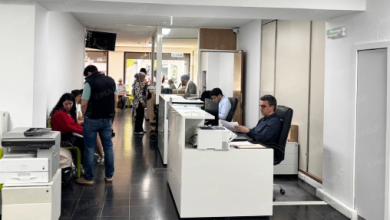 Open-plan office reception with white desks, staff at computers, and people seated or standing in a hallway waiting or talking.