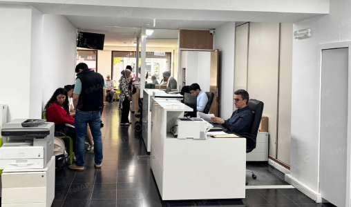 Open-plan office reception with white desks, staff at computers, and people seated or standing in a hallway waiting or talking.
