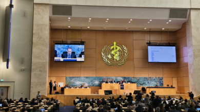 UN General Assembly chamber with a panel at the dais, audience seated, and the WHO emblem on the wall behind the speakers; two screens display a presenter.