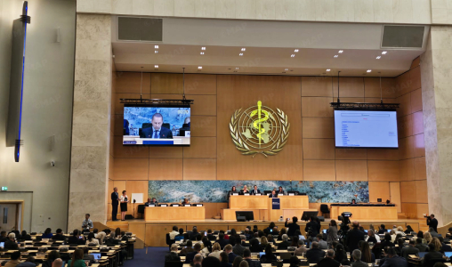 UN General Assembly chamber with a panel at the dais, audience seated, and the WHO emblem on the wall behind the speakers; two screens display a presenter.