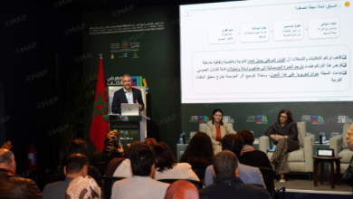 Speaker at a podium presenting at a conference; Moroccan flag behind, panelists seated on stage to the right, and an audience facing the stage with a large screen displaying Arabic text.