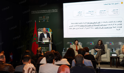 Speaker at a podium presenting at a conference; Moroccan flag behind, panelists seated on stage to the right, and an audience facing the stage with a large screen displaying Arabic text.
