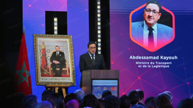 Speaker giving a formal address at a podium, with a framed portrait and Moroccan flag nearby; a large screen shows Abdessamad Kayouh and his title.