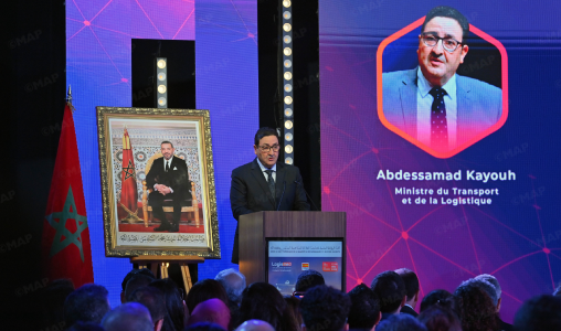 MAP54132134000 | MCG24 | موقع إلكتروني مغربي شامل متجدد على مدار الساعة Speaker giving a formal address at a podium, with a framed portrait and Moroccan flag nearby; a large screen shows Abdessamad Kayouh and his title.