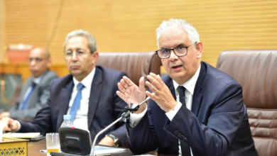 Three businessmen in a formal meeting; the man on the right speaks with hand gestures into a microphone.