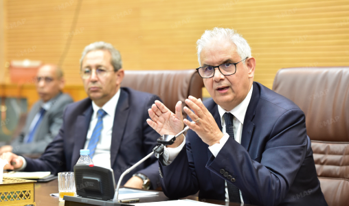 Three businessmen in a formal meeting; the man on the right speaks with hand gestures into a microphone.