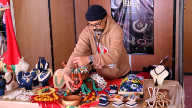 Vendor in a brown hoodie and beanie sorts beaded necklaces at a colorful jewelry stall on a wooden backdrop.
