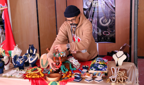 Vendor in a brown hoodie and beanie sorts beaded necklaces at a colorful jewelry stall on a wooden backdrop.