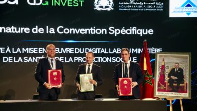 Three men in suits hold red and green folders on stage during a formal signing ceremony, Moroccan flag visible nearby.