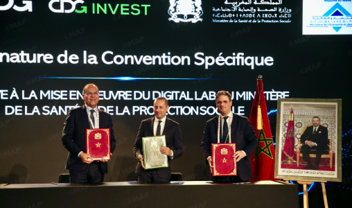 Three men in suits hold red and green folders on stage during a formal signing ceremony, Moroccan flag visible nearby.