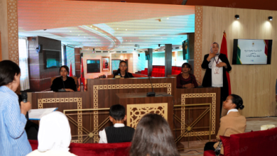 Panel discussion in a conference room with four panelists at a decorative wooden desk; a man speaks into a microphone at the right edge of the stage.