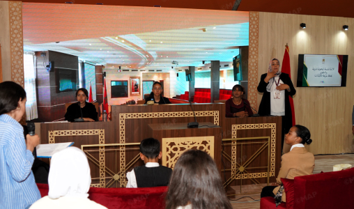 Panel discussion in a conference room with four panelists at a decorative wooden desk; a man speaks into a microphone at the right edge of the stage.