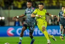 Two soccer players battle for the ball on a nighttime field; one in gray kit and one in neon yellow.