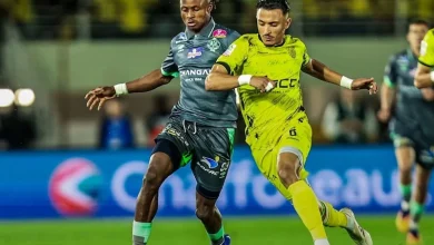 Two soccer players battle for the ball on a nighttime field; one in gray kit and one in neon yellow.