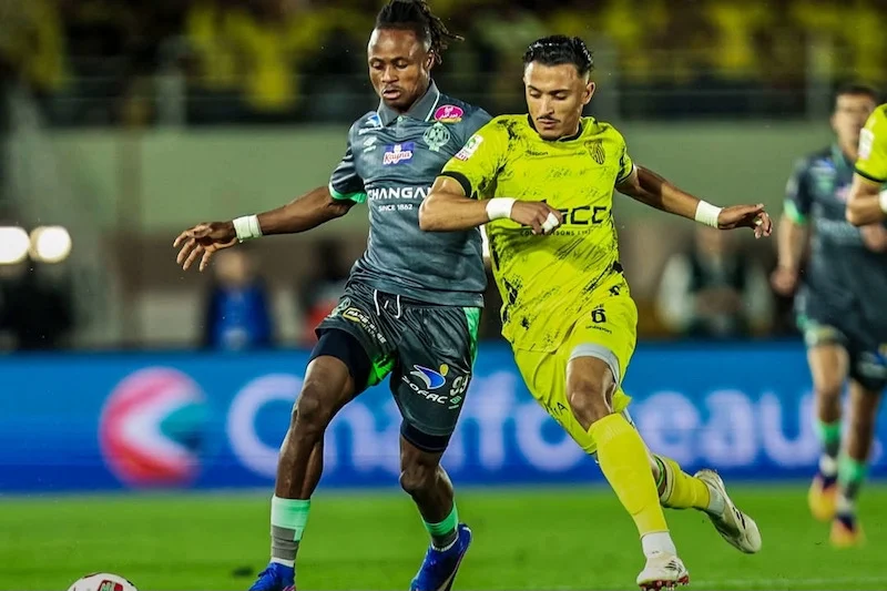 Two soccer players battle for the ball on a nighttime field; one in gray kit and one in neon yellow.