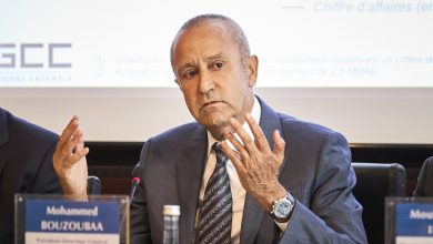 Man in a blue suit speaks at a conference, gesturing with both hands, with a microphone and nameplate in front reading 'Mohammad Bouzouba'.