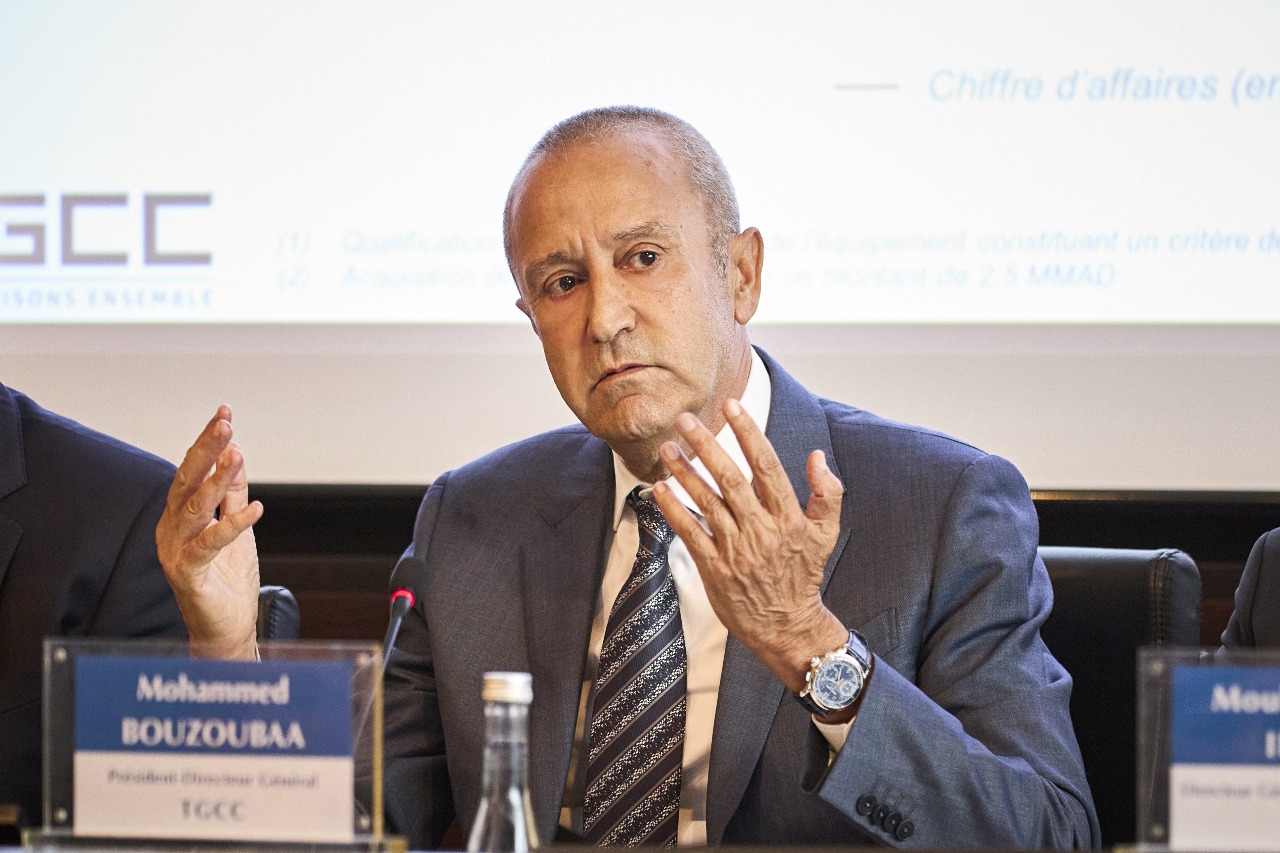 Man in a blue suit speaks at a conference, gesturing with both hands, with a microphone and nameplate in front reading 'Mohammad Bouzouba'.